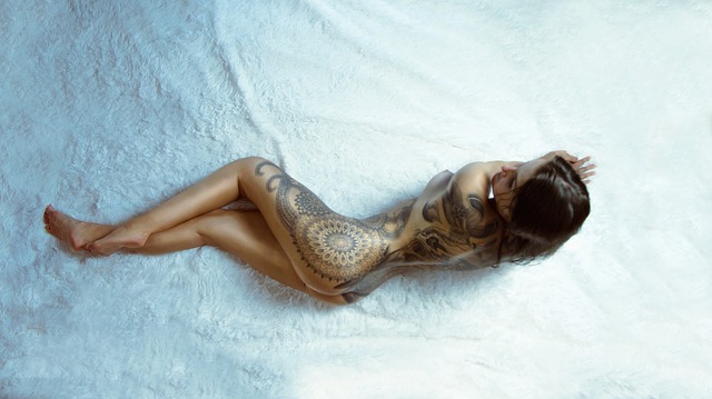 naked tattoed woman lying on white sheets of a large bed